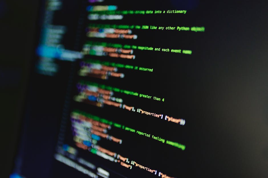 Close-up view of a computer screen displaying lines of colorful programming code with a black background. The code includes text in green, orange, blue, and white, with parts of it slightly blurred du