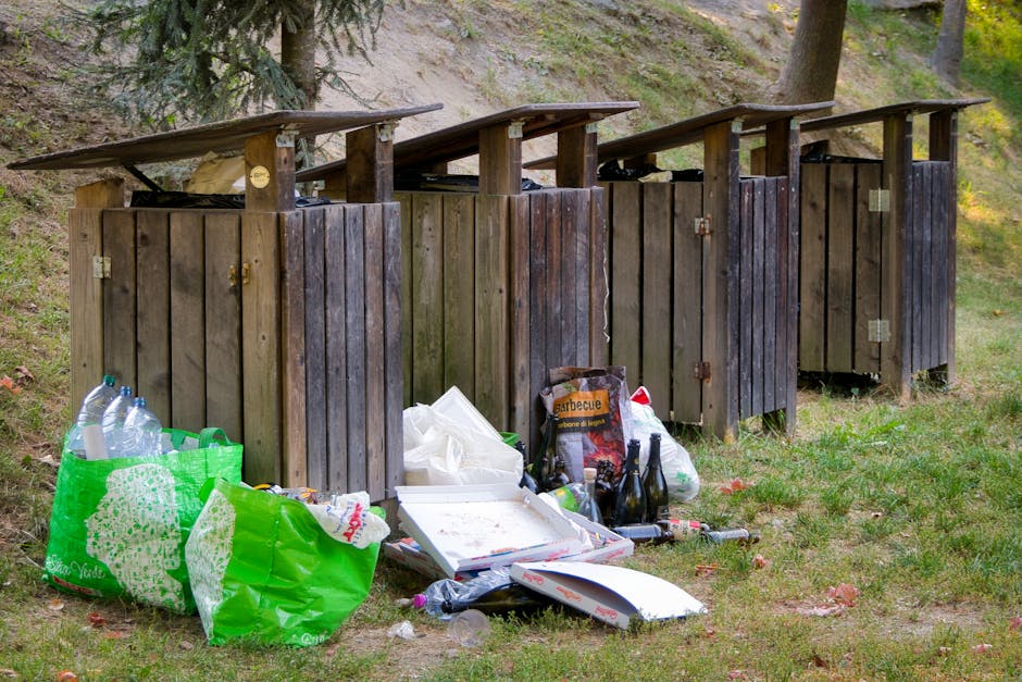 Four wooden outdoor rubbish bins with slanted flat roofs are arranged side by side on a grassy area near trees. The bins are weathered, with darkened wood panels showing signs of use and age. In front